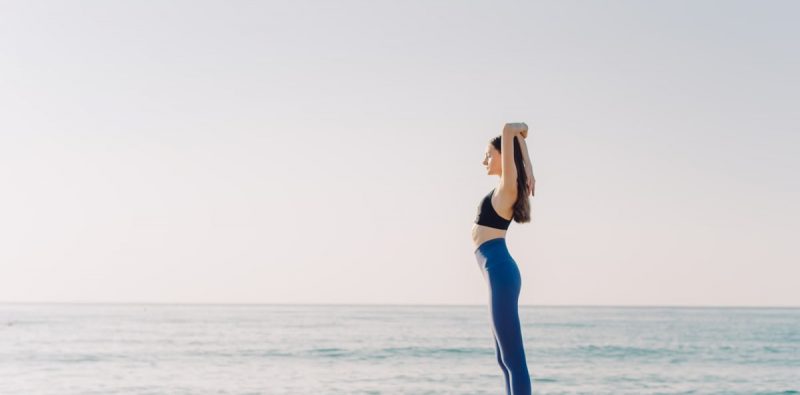 A woman performing yoga on a serene beach with a clear blue sky, emphasizing relaxation and wellness.